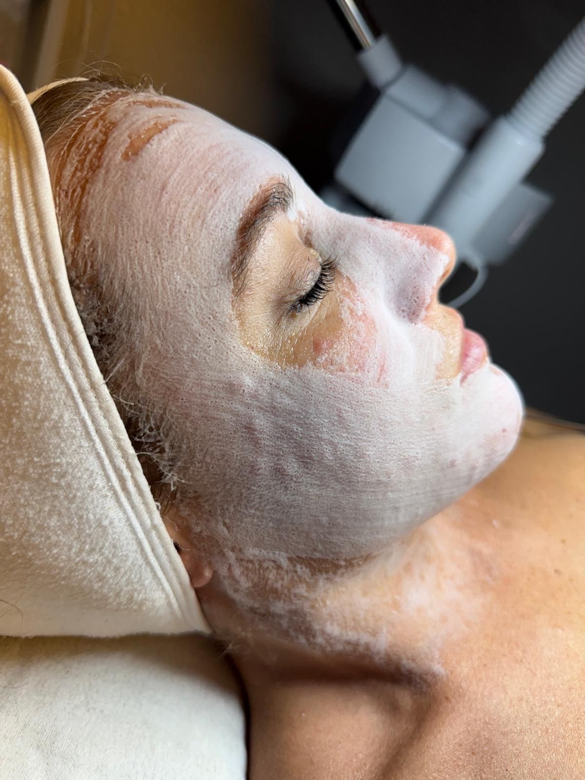 Mujer recibiendo un tratamiento facial con una mascarilla blanca aplicada, tumbada en una camilla de spa.
