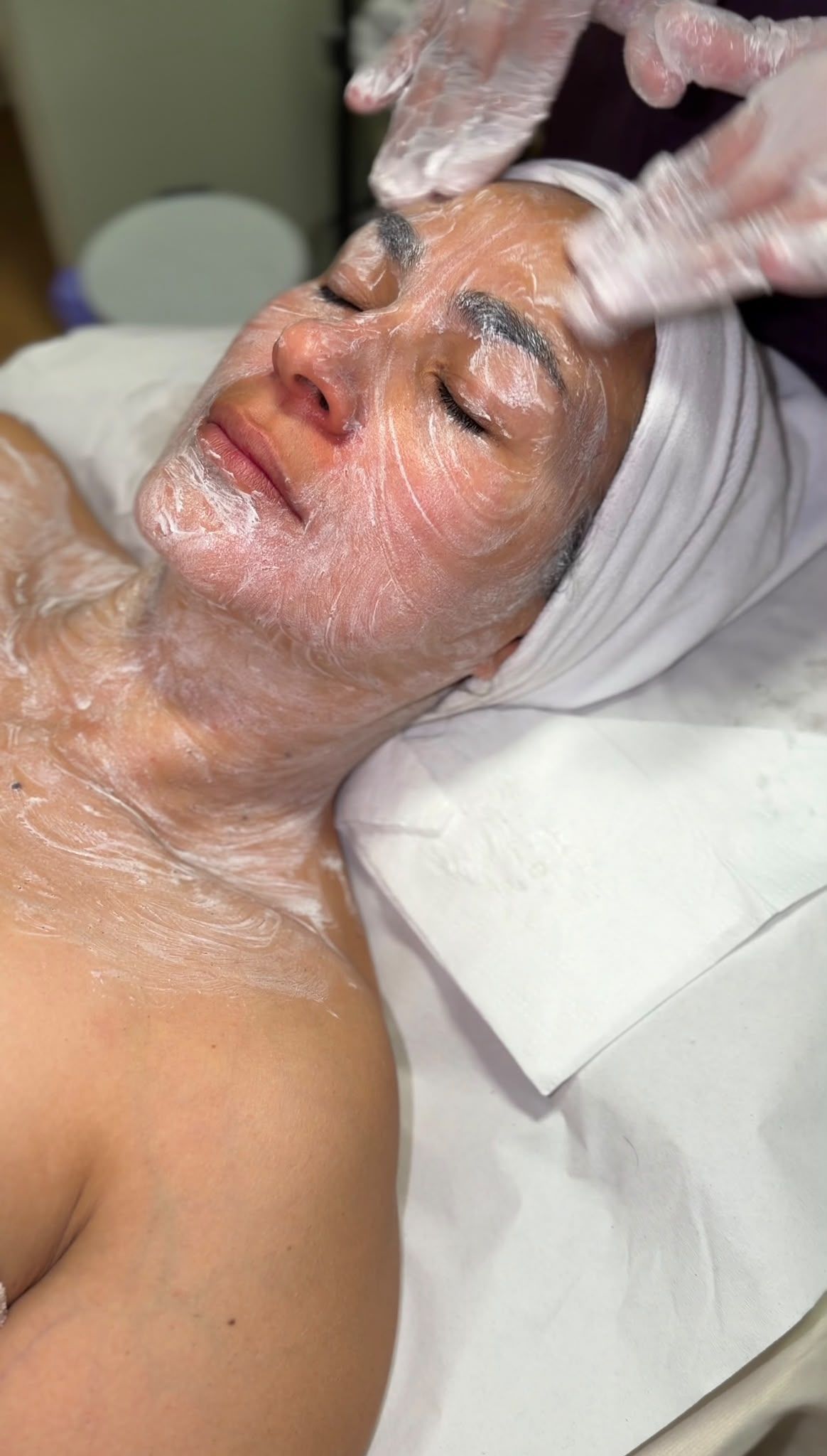 Persona recibiendo un tratamiento facial con crema blanca en la cara y el cuello, tumbada en una camilla de spa.