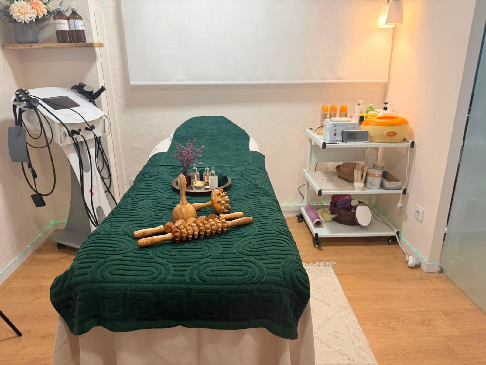 Camilla de masaje con manta verde, toallas y carrito con ruedas en una sala de tratamientos de spa.