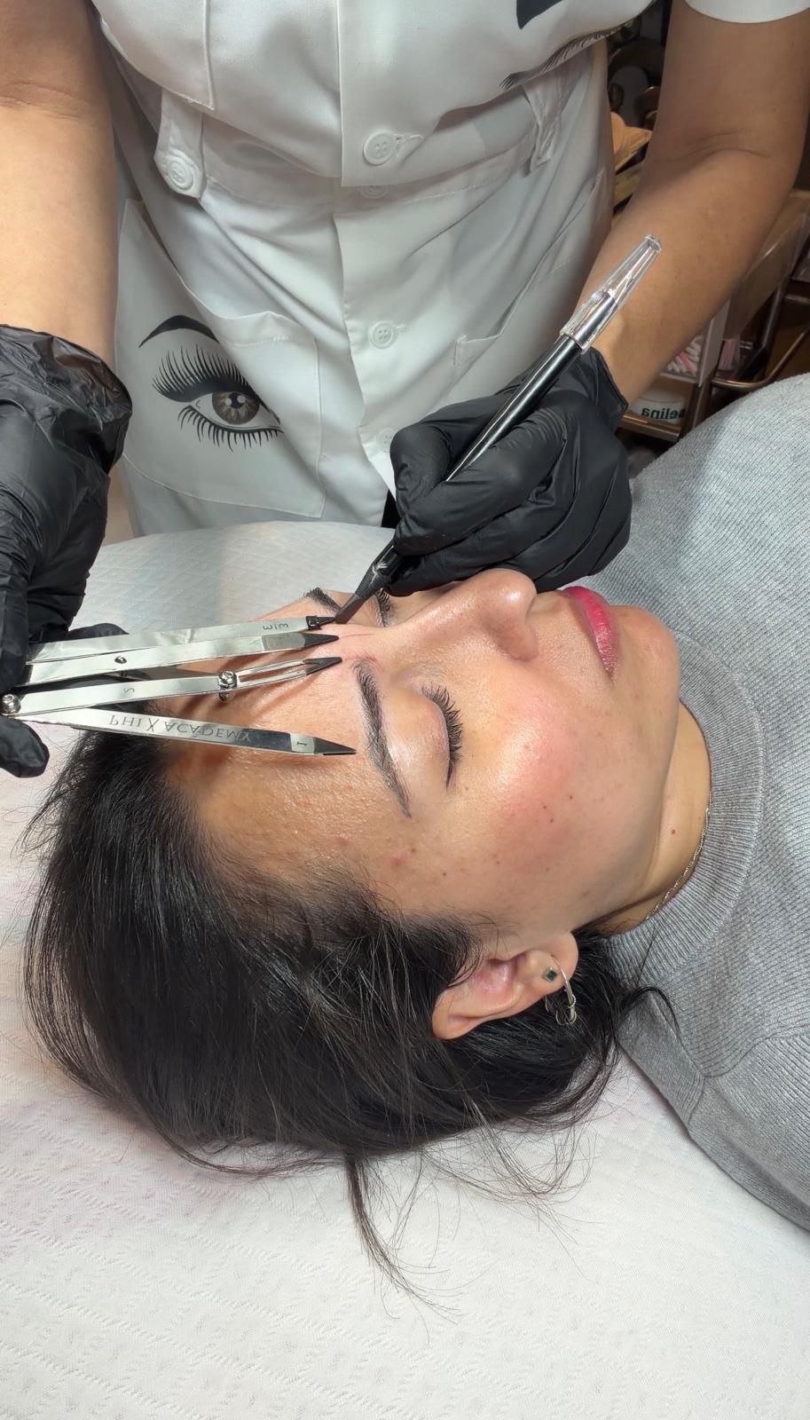 Esteticista aplica un tratamiento de cejas con un pincel a una clienta tumbada en una camilla de tratamiento.