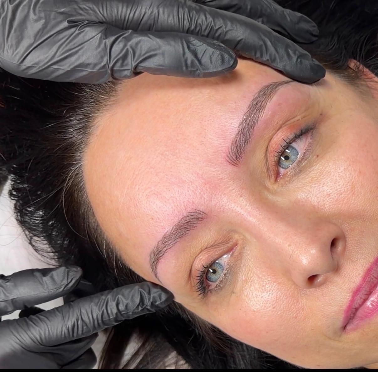 Manos enguantadas sosteniendo el rostro de una mujer durante un tratamiento cosmético de cejas.