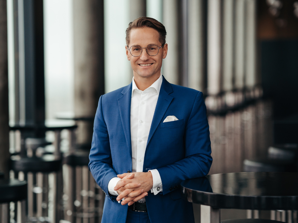 Ein Mann im blauen Anzug lächelt und lehnt sich an den Tisch. Weißes Einstecktuch, Brille, formelles Ambiente.