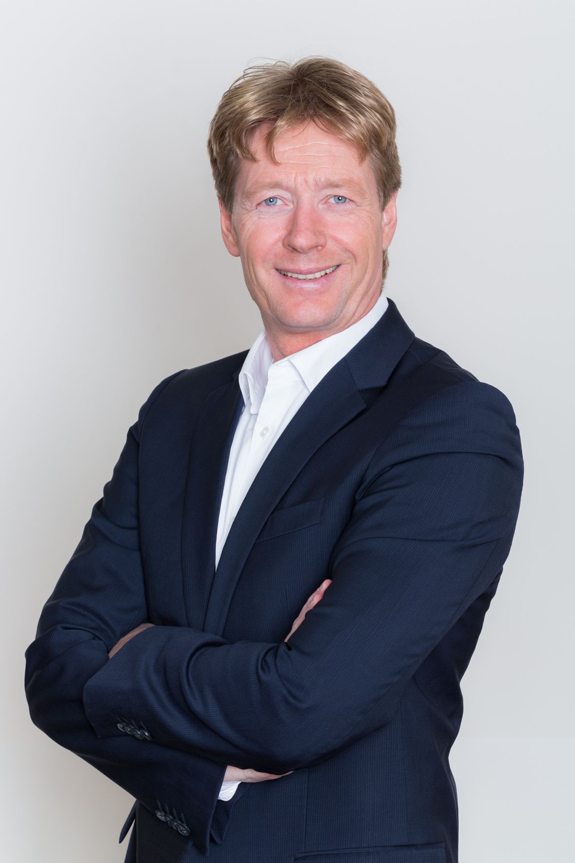 Mann mit blonden Haaren, in einem dunkelblauen Blazer und weißem Hemd, die Arme verschränkt, lächelnd.