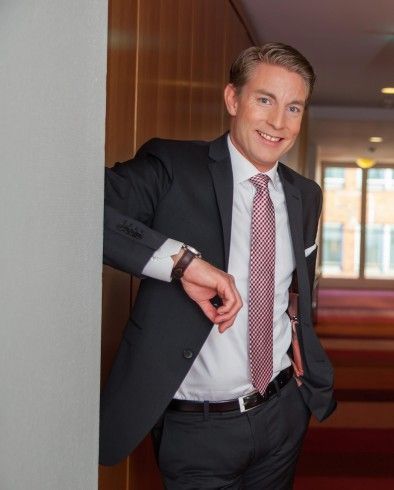 Ein Mann im Anzug lächelt und lehnt drinnen an einer Wand. Er trägt Krawatte, Uhr und Gürtel.