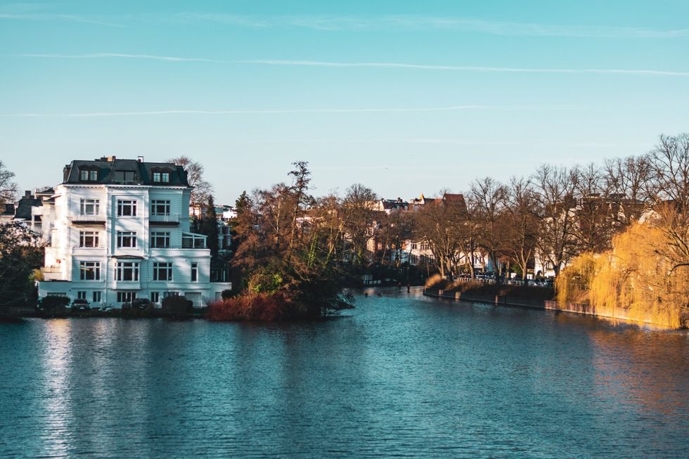 Weißes mehrstöckiges Haus an einem See mit Bäumen unter blauem Himmel.