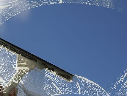 Une raclette essuie la mousse savonneuse d'une vitre, révélant un ciel bleu limpide.