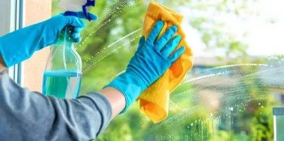 Une personne portant des gants bleus essuie une vitre avec un chiffon jaune tout en tenant un flacon pulvérisateur.