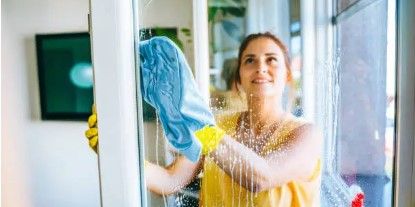 Une personne vêtue d'un haut jaune et portant des gants de nettoyage jaunes essuie une vitre avec un chiffon bleu.