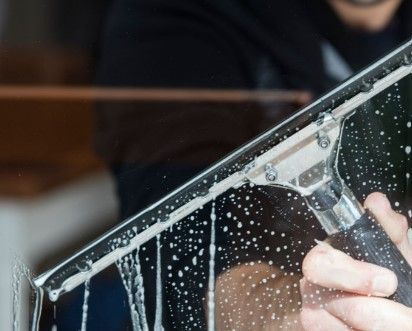 Une personne nettoie une vitre savonneuse avec une raclette métallique, laissant une trace transparente en essuyant la mousse.