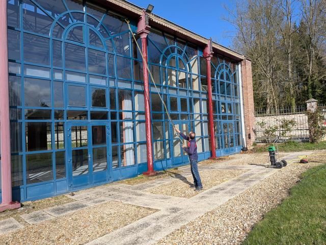 Une personne nettoie de grandes fenêtres cintrées à cadre bleu sur un bâtiment en briques à l'aide d'une brosse à manche allongé.