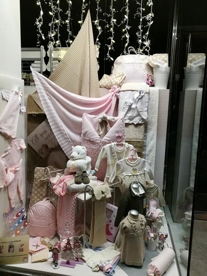 Escaparate de ropa de bebé en tonos rosas y cremas, con vela decorativa y guirnalda de luces.