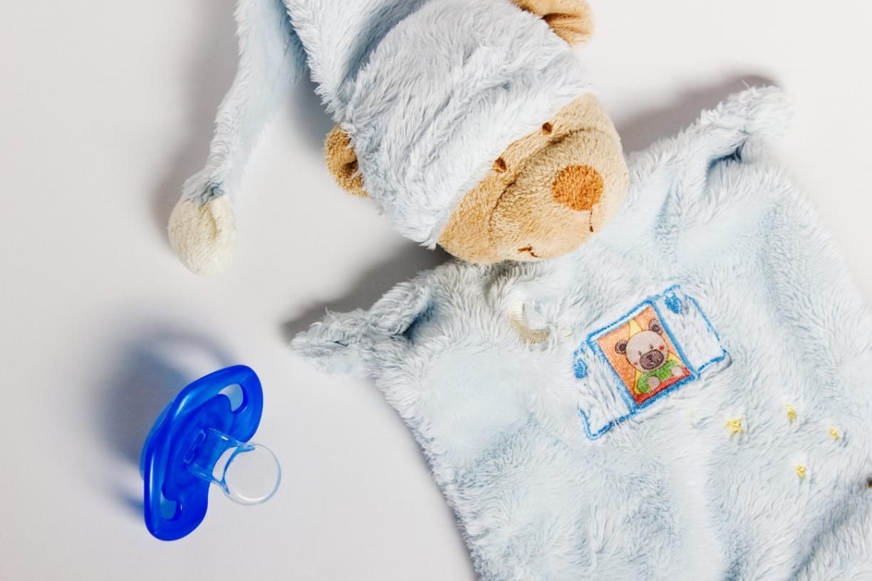 Chupete azul, gorro de osito de peluche y mono azul peludo sobre superficie blanca.