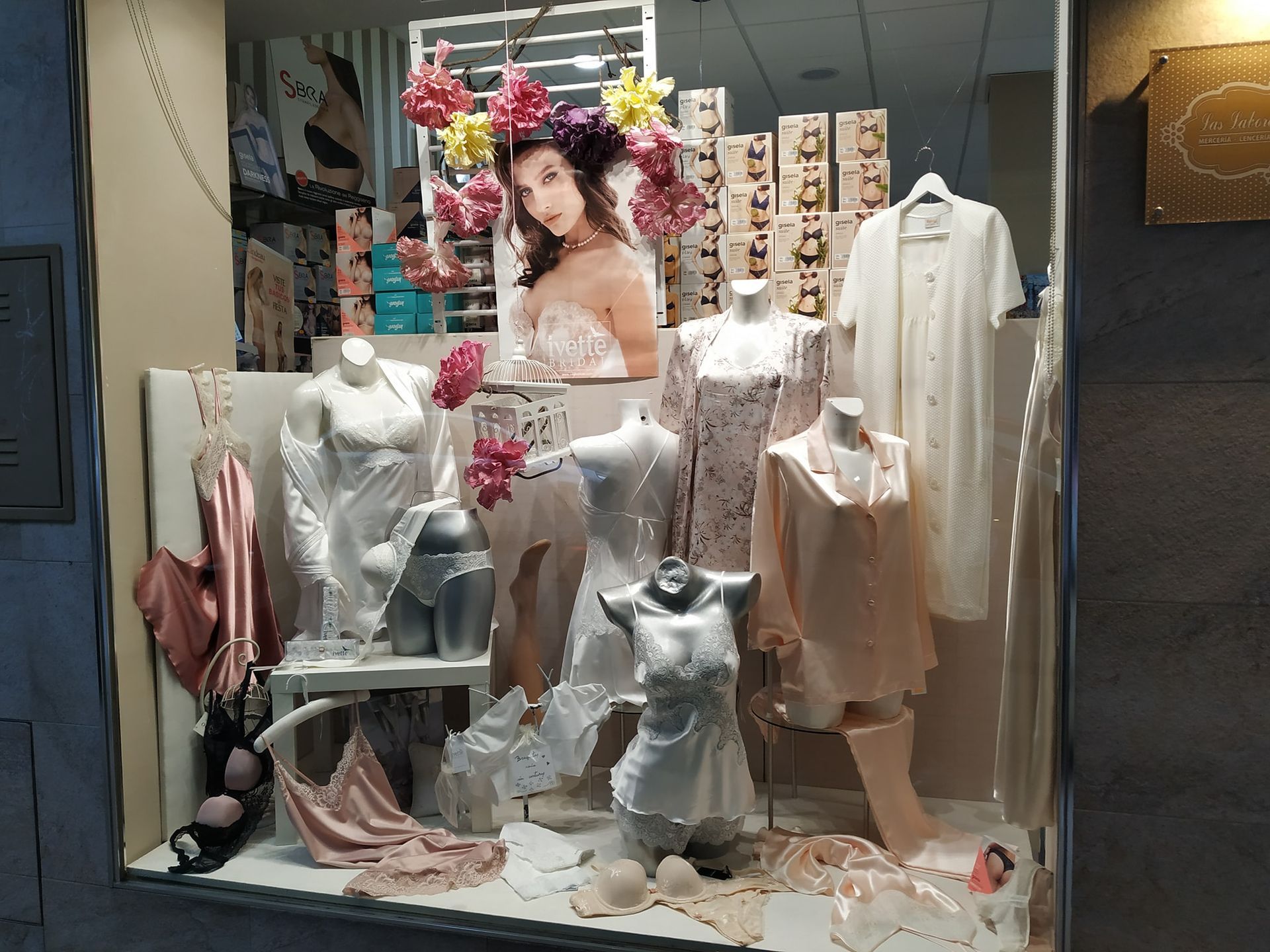 Exhibición de tienda de lencería con maniquíes vistiendo ropa de dormir blanca y rosa y una mujer con flores, una ventana.