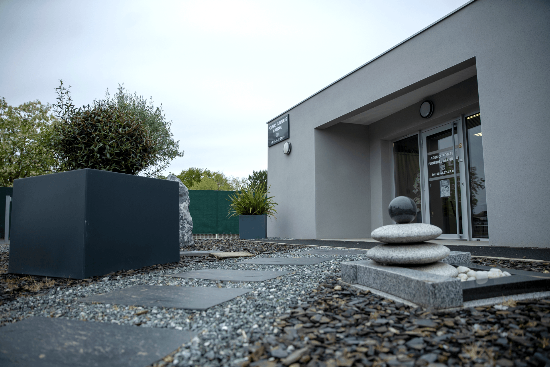 Extérieur d'un bâtiment moderne avec jardin zen. Murs gris, allée de pierre, jardinières sombres, pierres empilées.