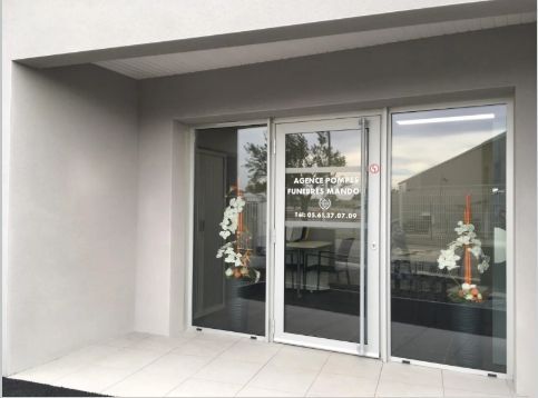 Extérieur d'une maison funéraire avec portes vitrées, fleurs et enseigne. Bâtiment gris, boiseries blanches.