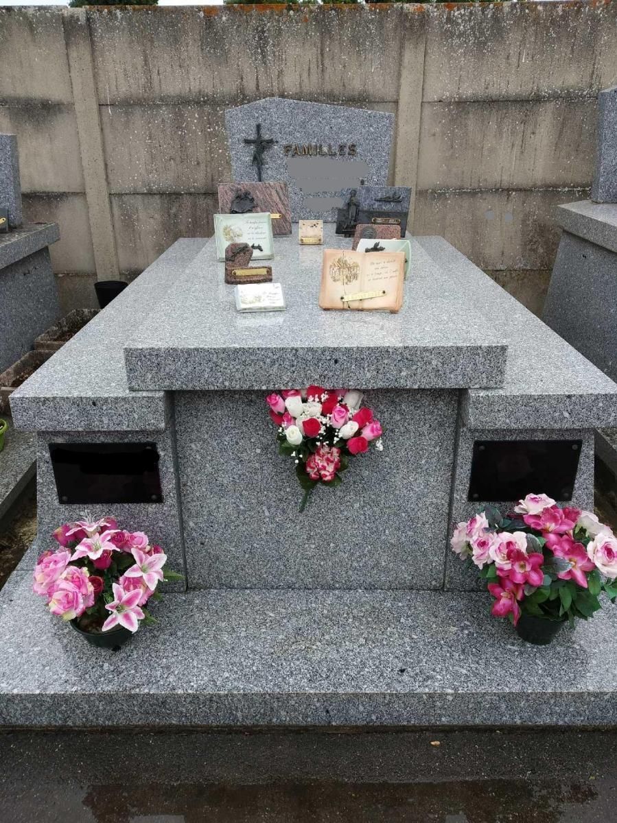 Pierre tombale en granit gris ornée de fleurs et surmontée de petits objets religieux, dans un cimetière.