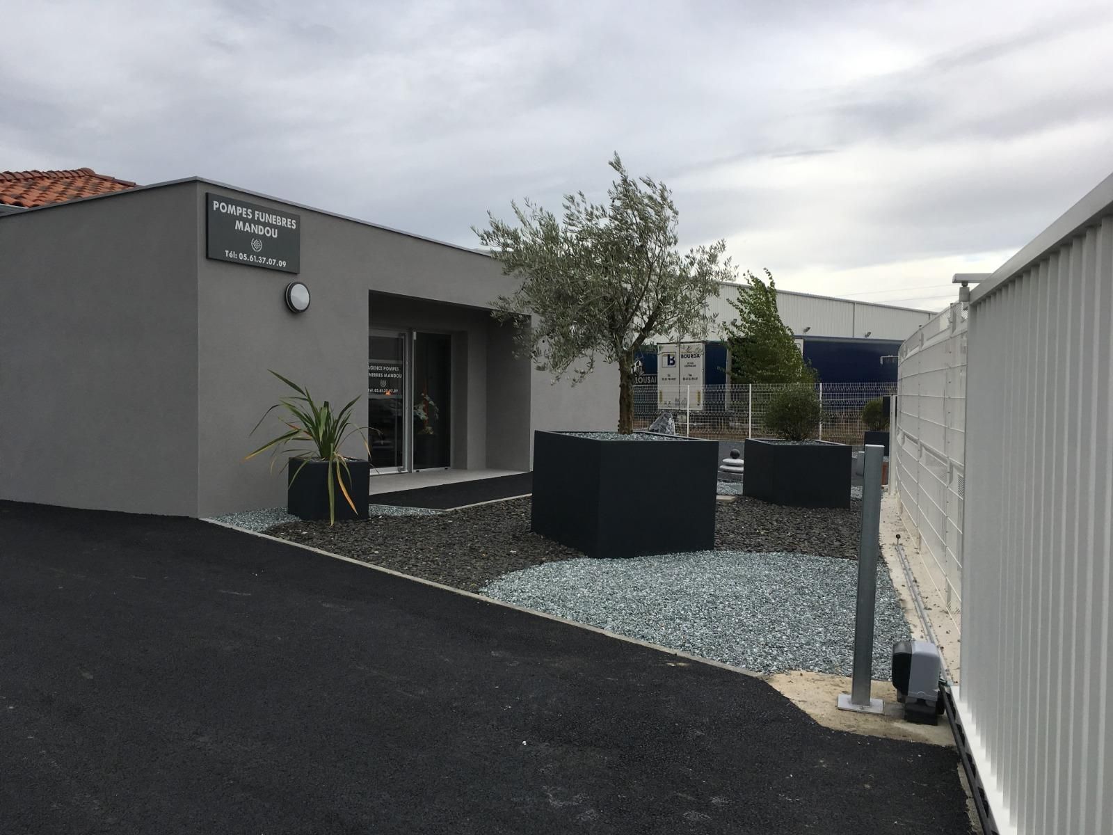 Bâtiment gris avec enseigne, petits arbres dans des jardinières noires et gravier foncé.