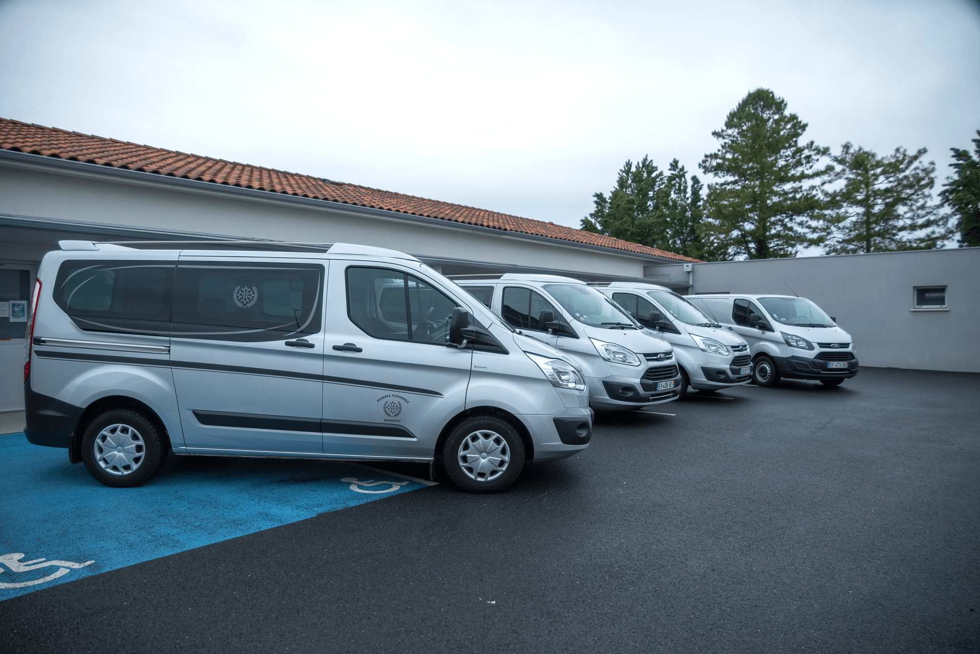 Cinq fourgonnettes argentées sont stationnées devant un bâtiment, une zone d'accessibilité étant peinte en bleu. Ciel couvert.