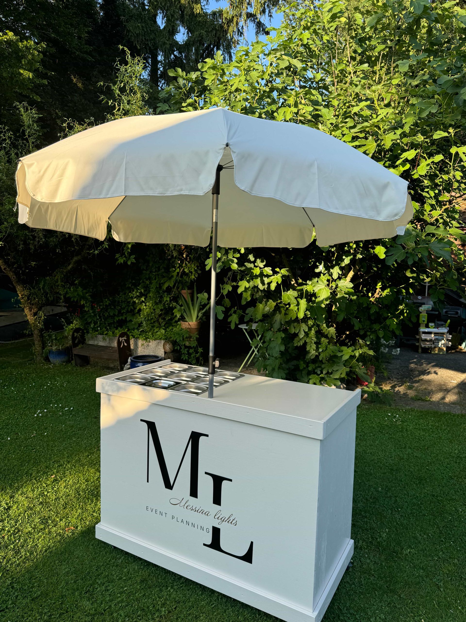 Deux femmes devant un stand événementiel. L'une prépare à manger, l'autre observe. Le stand est blanc et porte le logo « ML ».