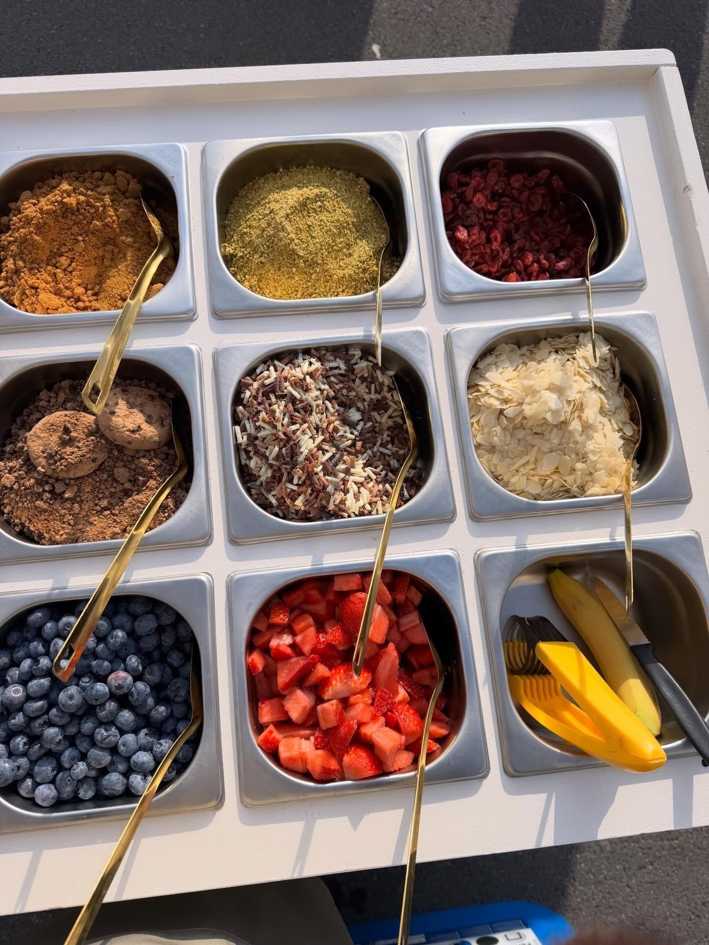 Un plateau de garnitures : granola, noix, baies, chocolat et fruits, avec des cuillères de service dorées.