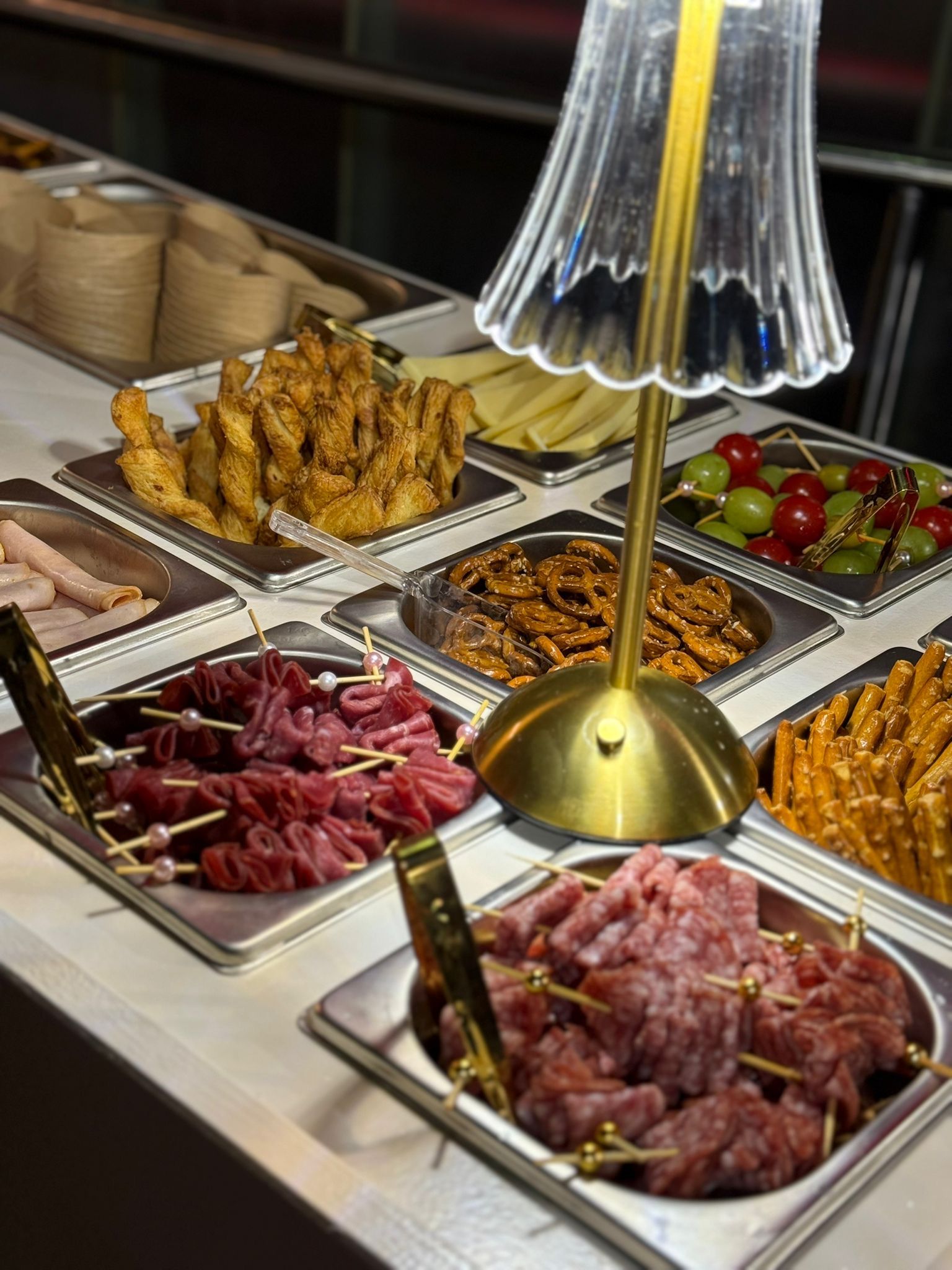 Une main tient un gobelet rempli de prosciutto à côté d'une table garnie d'amuse-gueules variés et d'une petite lampe.