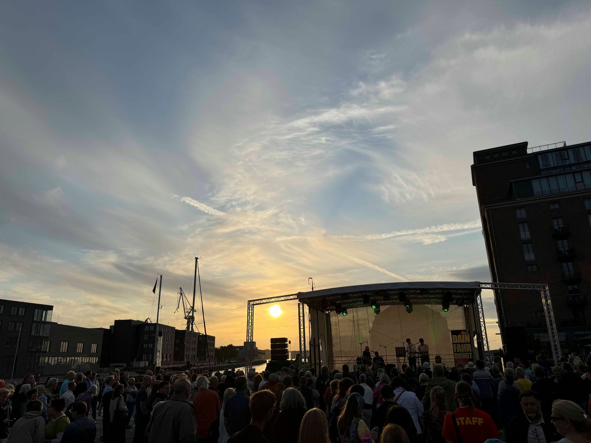 Konzert auf einer Bühne mit Publikum bei Sonnenuntergang, im Hintergrund Gebäude.
