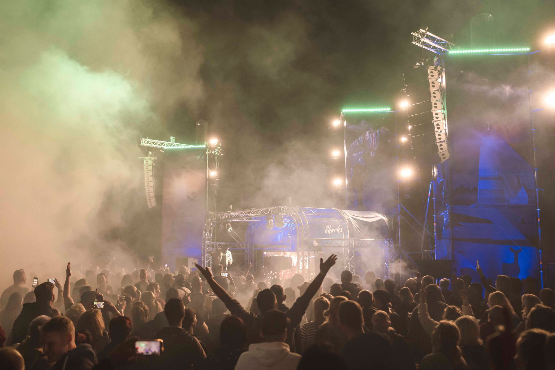 Eine Menschenmenge bei einem Konzert mit erhobenen Armen, die Bühne ist grün beleuchtet, Rauch liegt in der Luft.