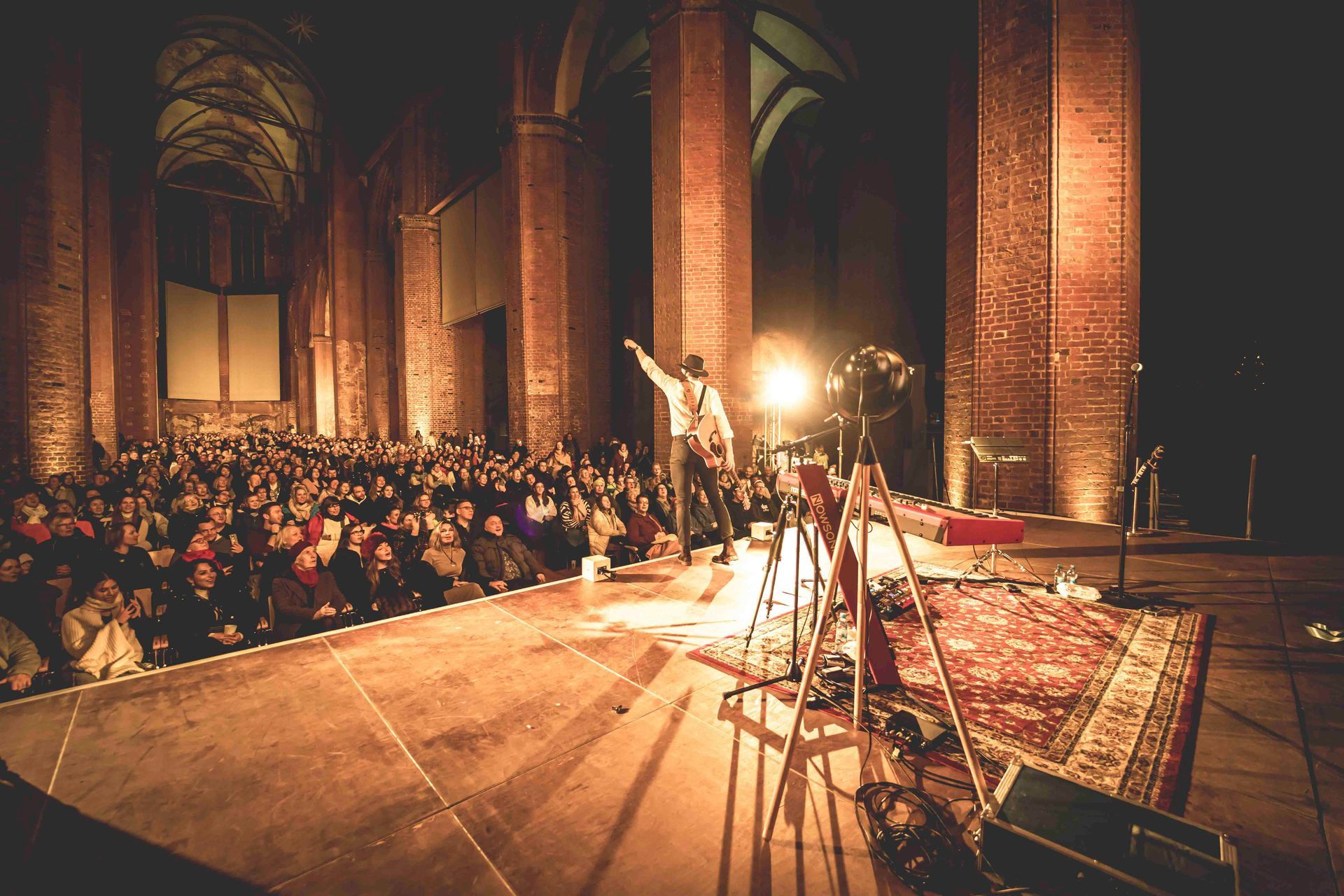 Ein Musiker tritt in einer großen, dunklen Halle vor einem sitzenden Publikum auf.