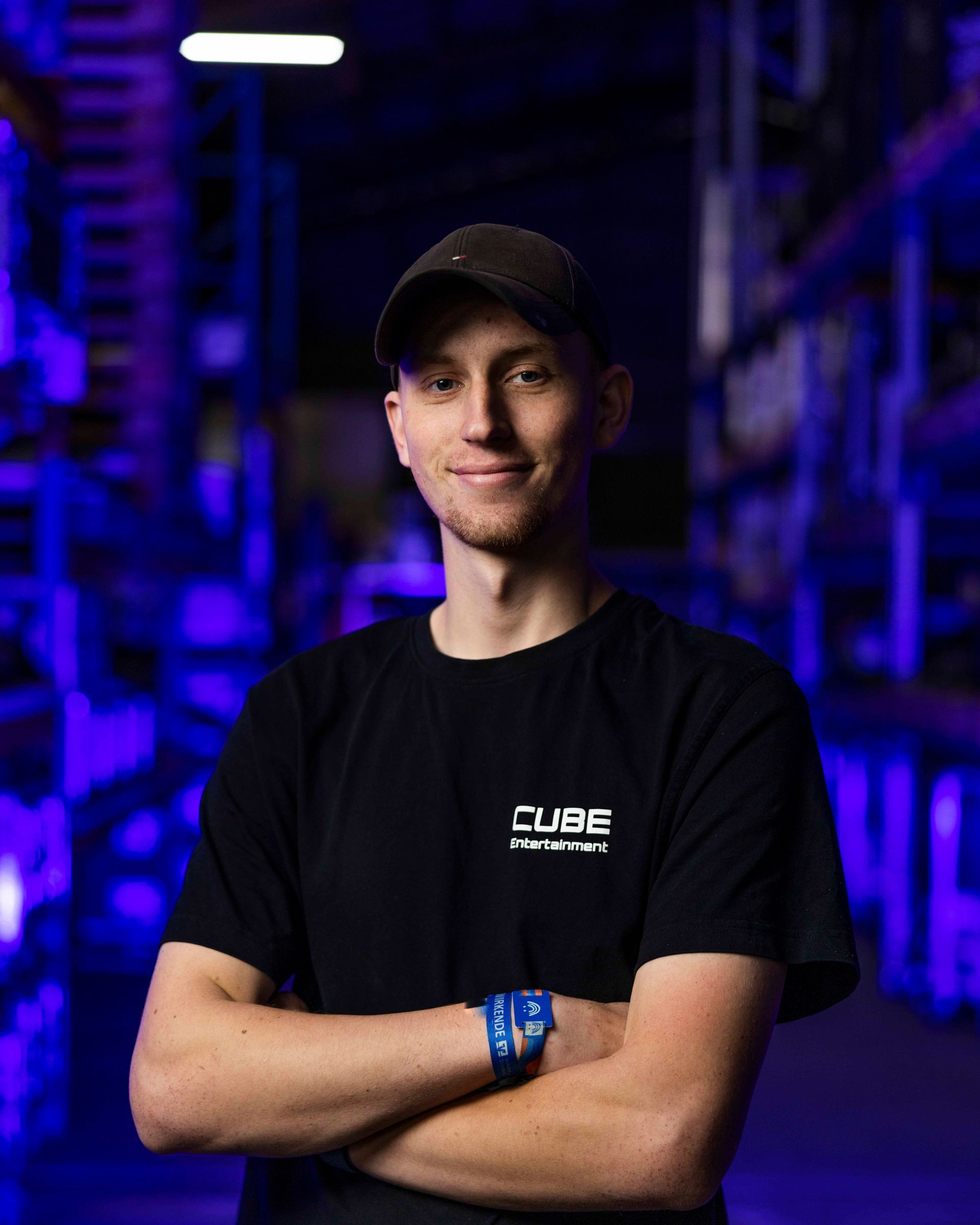 Mann in schwarzem T-Shirt und Kappe, die Arme verschränkt, lächelnd. Lagerhalle, blau-violette Beleuchtung.