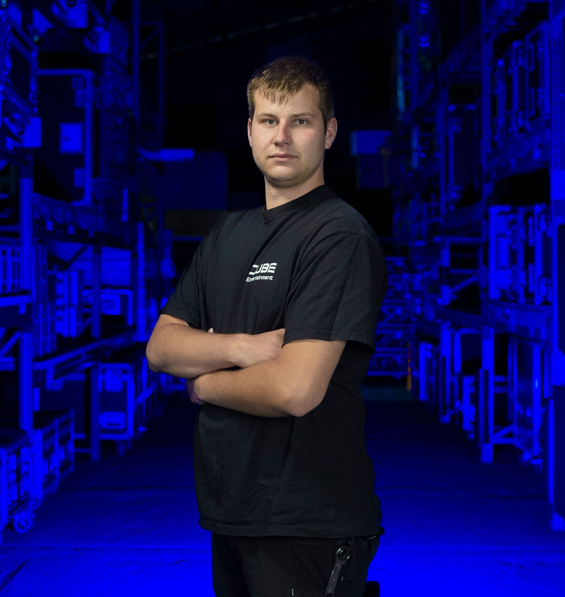 Mann in schwarzem Hemd mit verschränkten Armen steht in einer blau beleuchteten Lagerhalle.