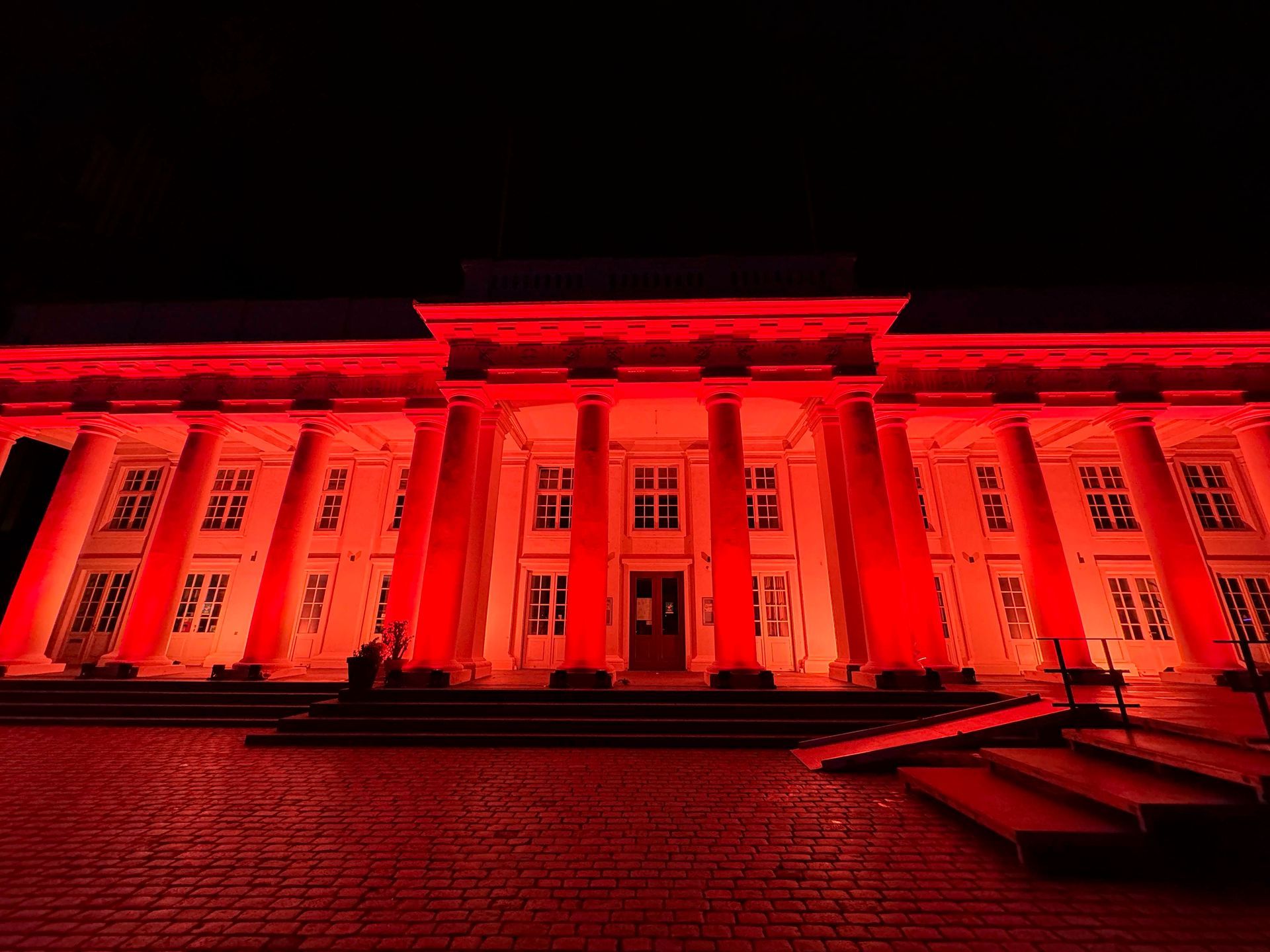 Ein neoklassizistisches Gebäude, nachts rot beleuchtet. Große Säulen und Fenster. Dunkler Himmel.