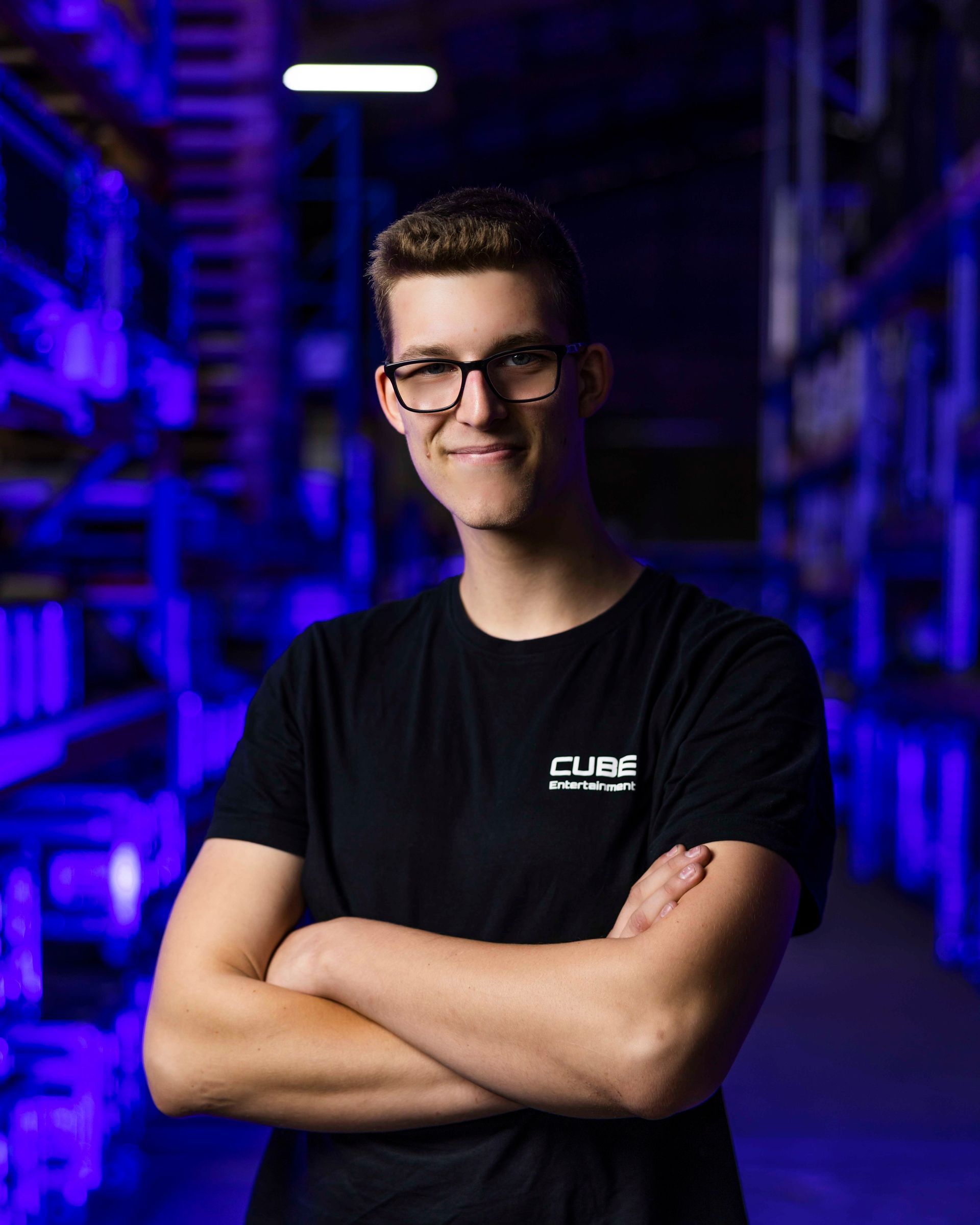 Mann mit Brille und verschränkten Armen, der ein schwarzes Hemd mit Logo trägt, in einer blau beleuchteten Lagerhalle.