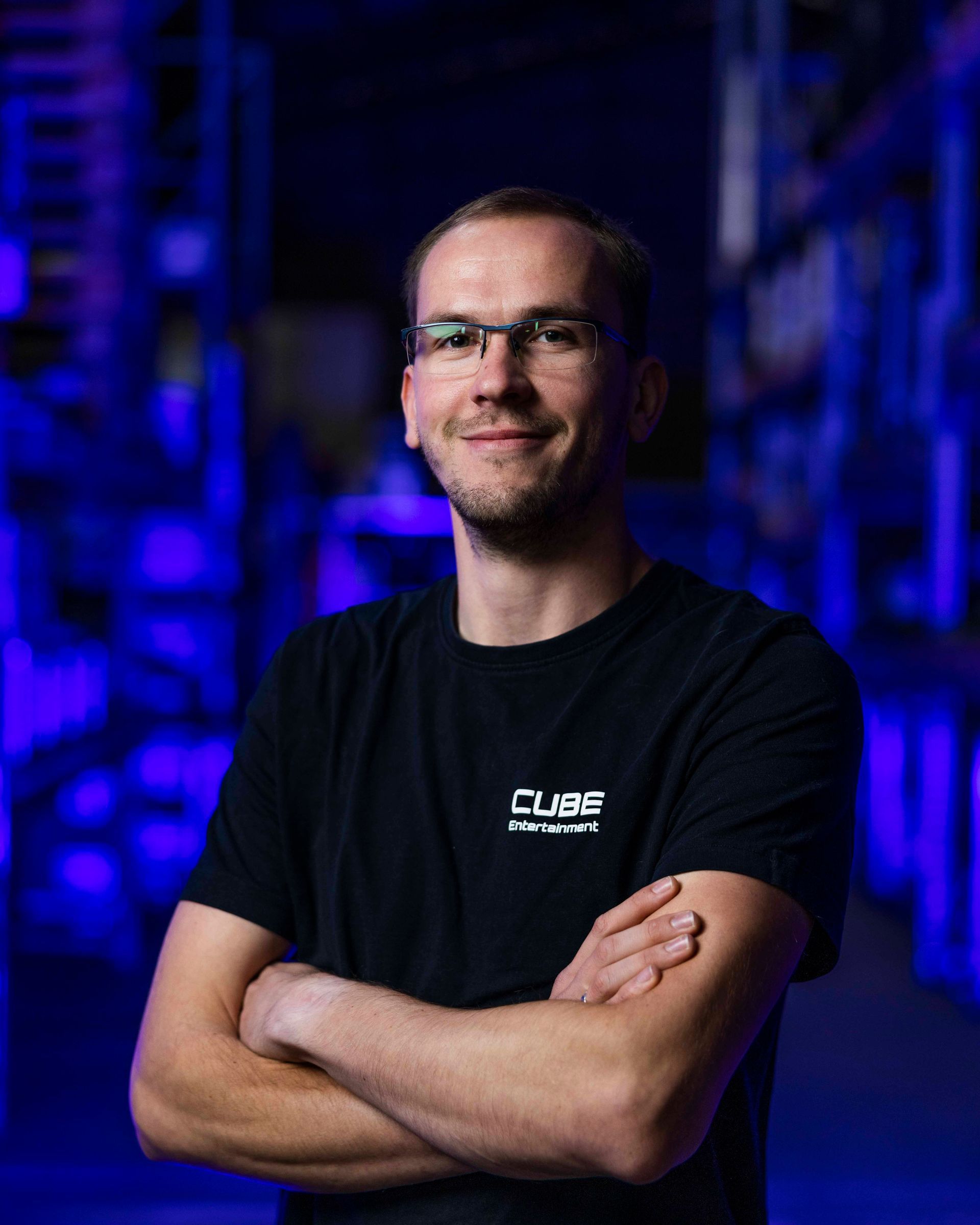 Mann mit Brille und schwarzem T-Shirt, die Arme verschränkt, lächelt. Verschwommener blauer Hintergrund.