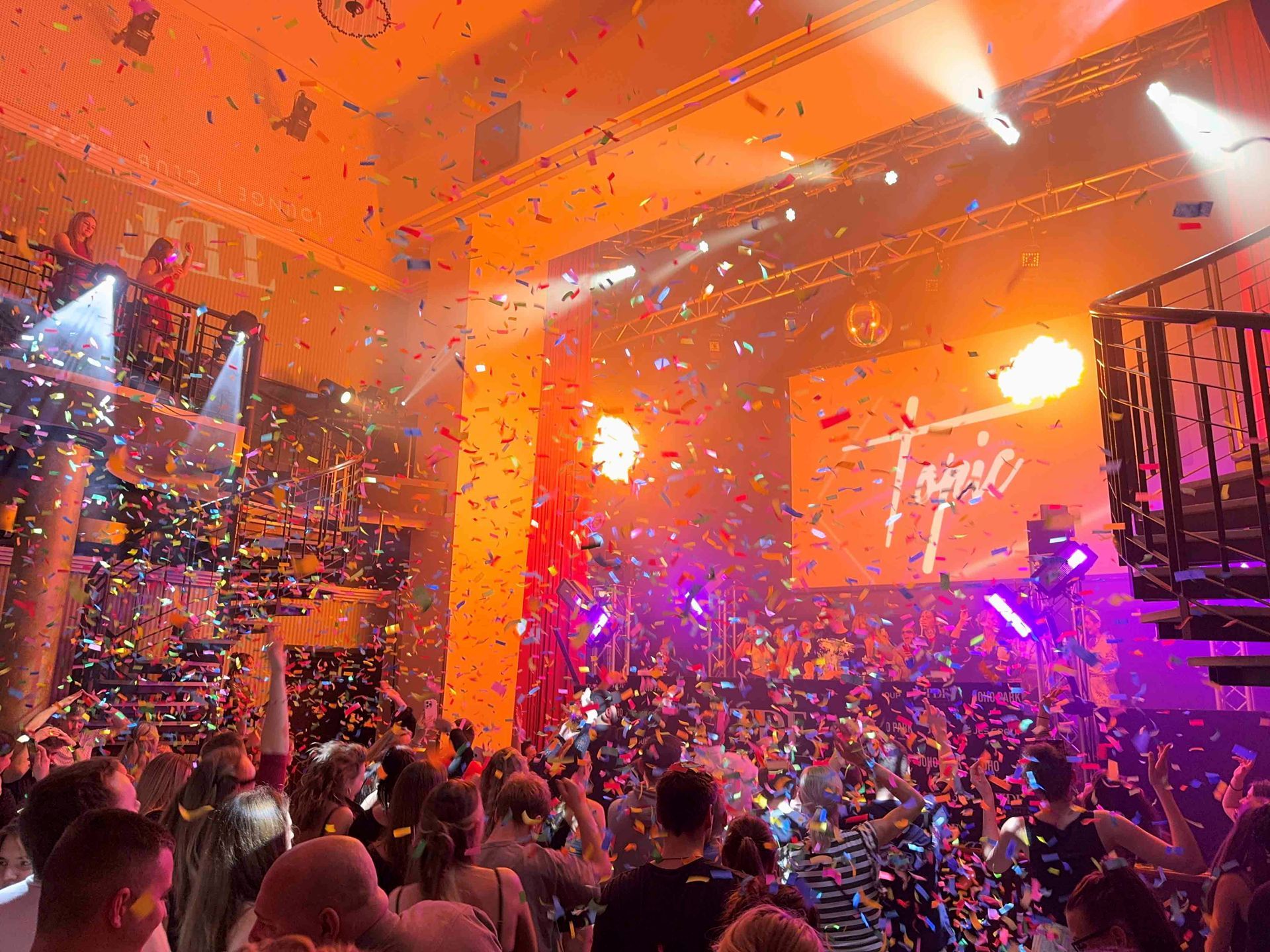 Eine mit Konfetti gefüllte Tanzfläche in einem Club; Menschen tanzen unter hellem Licht und vor einer Bühne mit Leinwand.