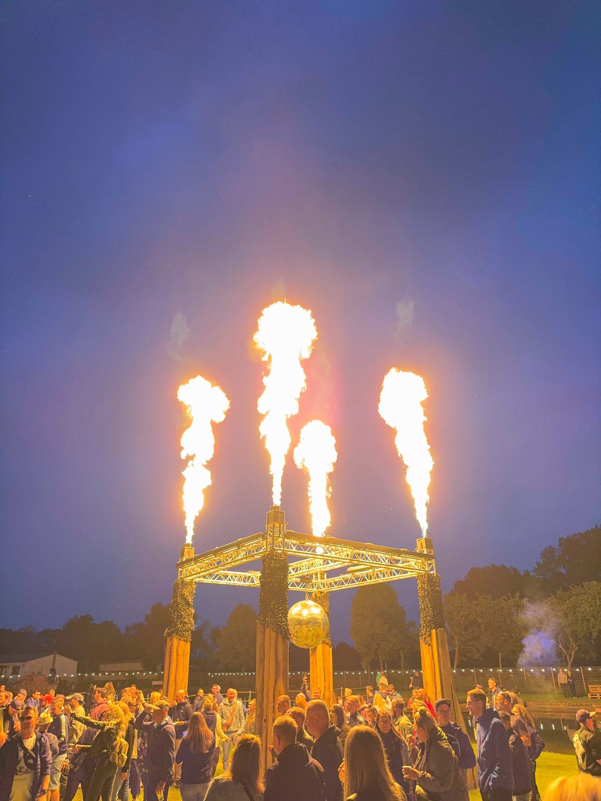 Während einer nächtlichen Veranstaltung schlagen Flammen aus einem Gebäude hervor; im Vordergrund beobachten Menschen das Geschehen.