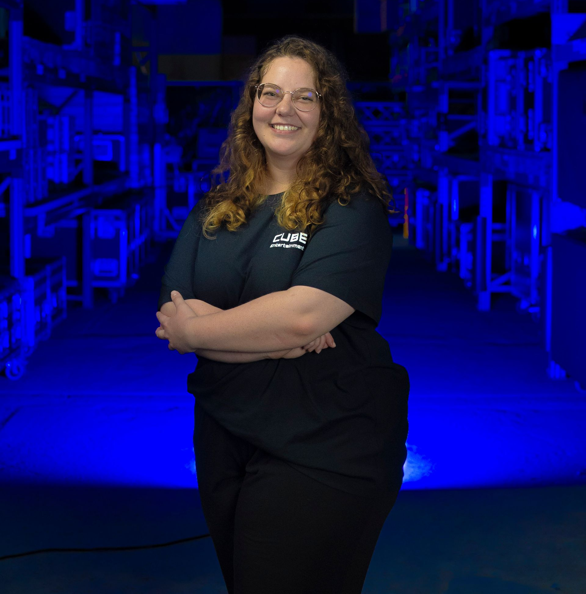 Frau mit Brille, verschränkten Armen, lächelnd. Sie trägt ein schwarzes Hemd und eine schwarze Hose und steht in einem blau beleuchteten Raum.