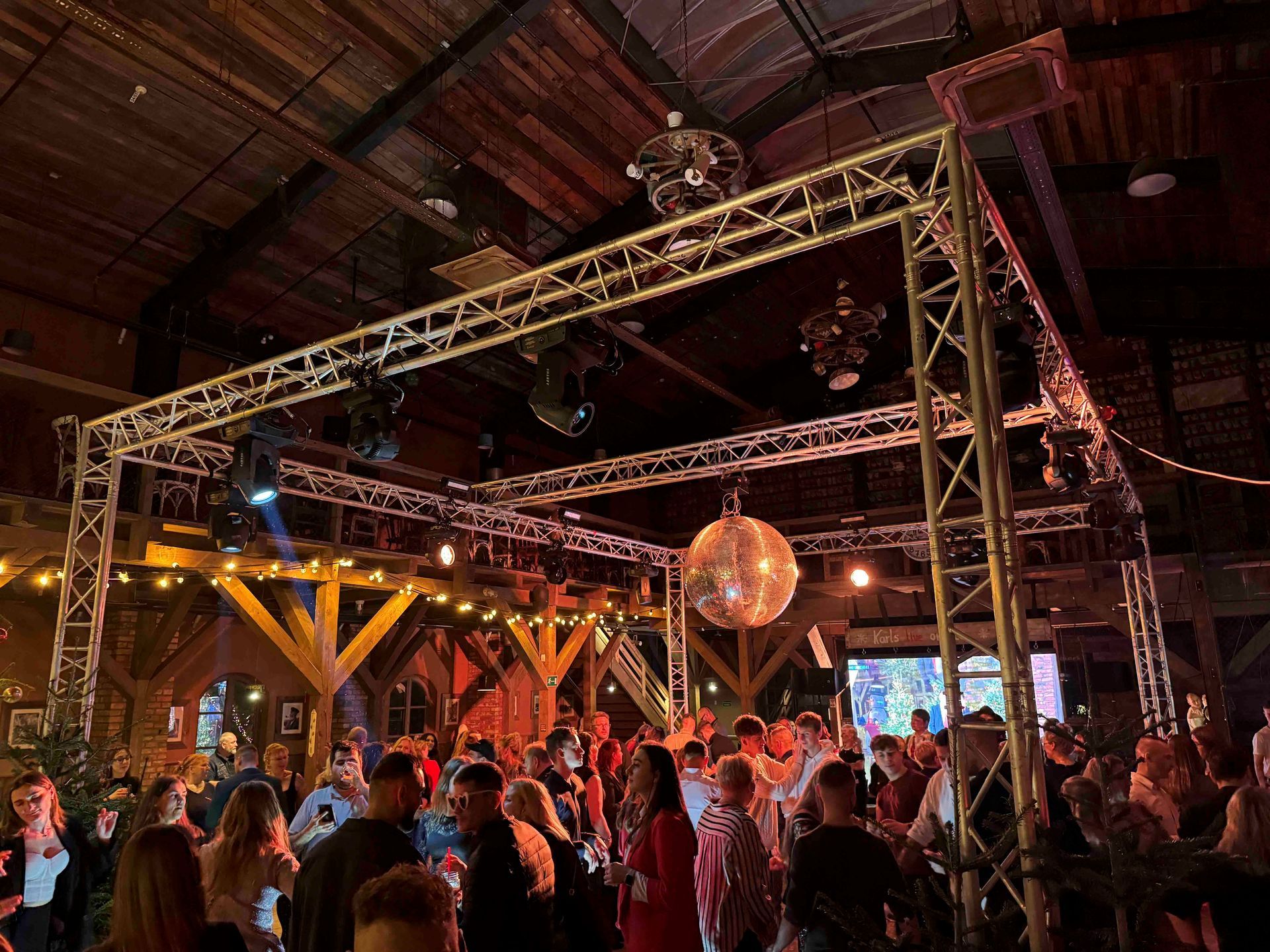 Party in einem rustikalen Saal: tanzende Menschen, Discokugeln, ein mit Lichtern bestücktes Metallgerüst und Holzbalken.
