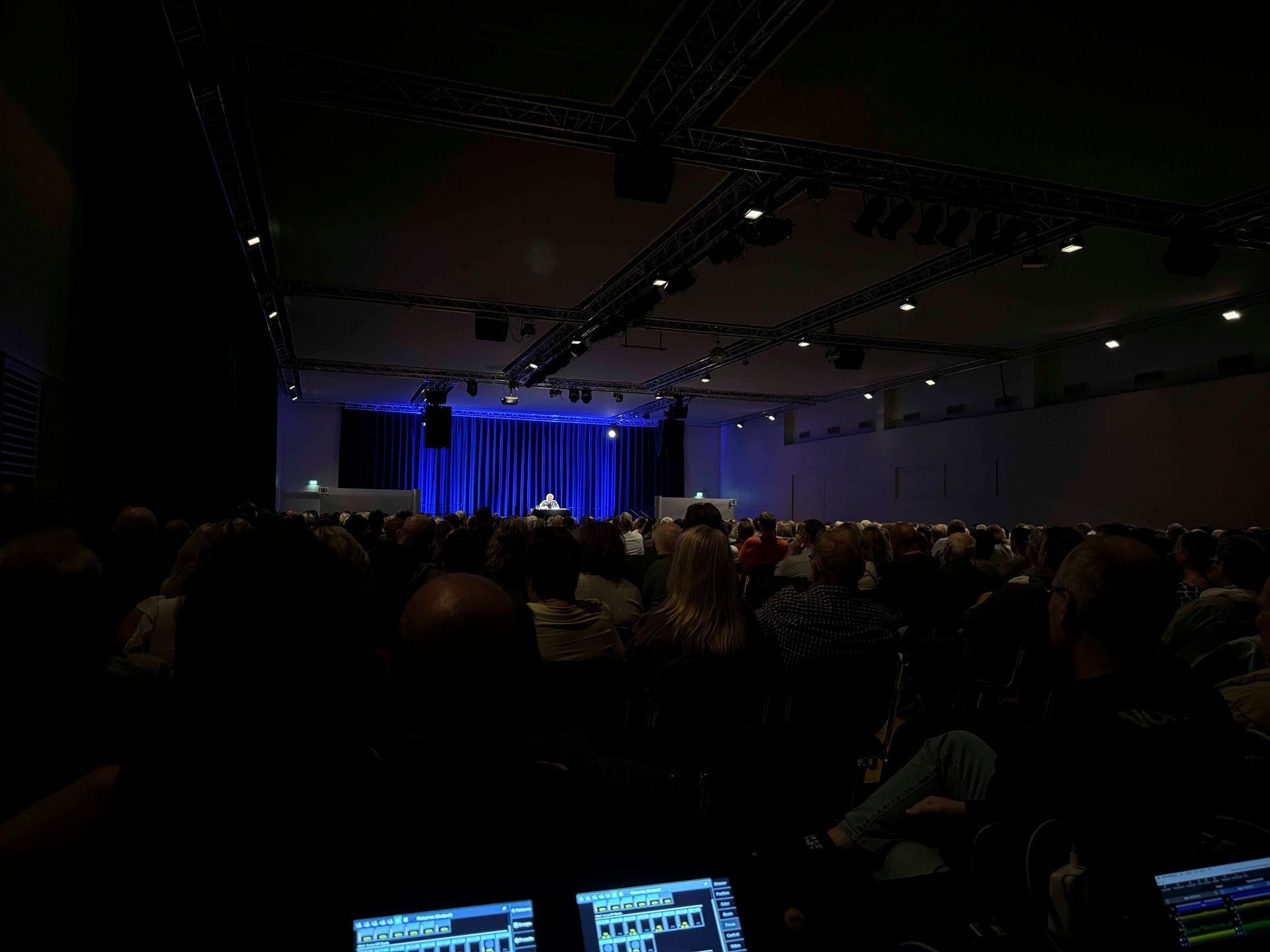Zuschauer in einem schwach beleuchteten großen Saal verfolgen eine Präsentation auf einer Bühne mit blauen Vorhängen und Scheinwerfern.