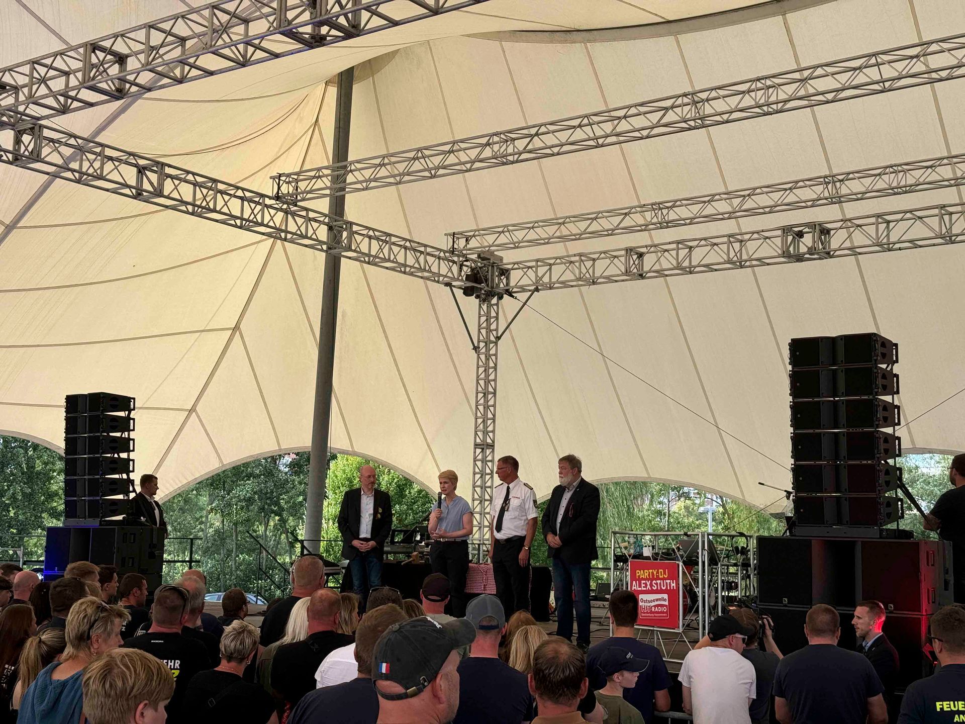 Menschen auf der Bühne bei einer Open-Air-Veranstaltung, davor Tonanlage und Publikum.