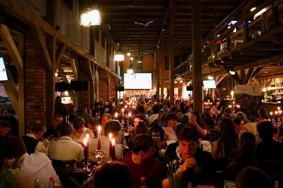 Große, stimmungsvoll beleuchtete Veranstaltung. Menschen sitzen an Tischen, viele Kerzen brennen. Holzbalken und Backsteinwände schaffen eine rustikale Atmosphäre.