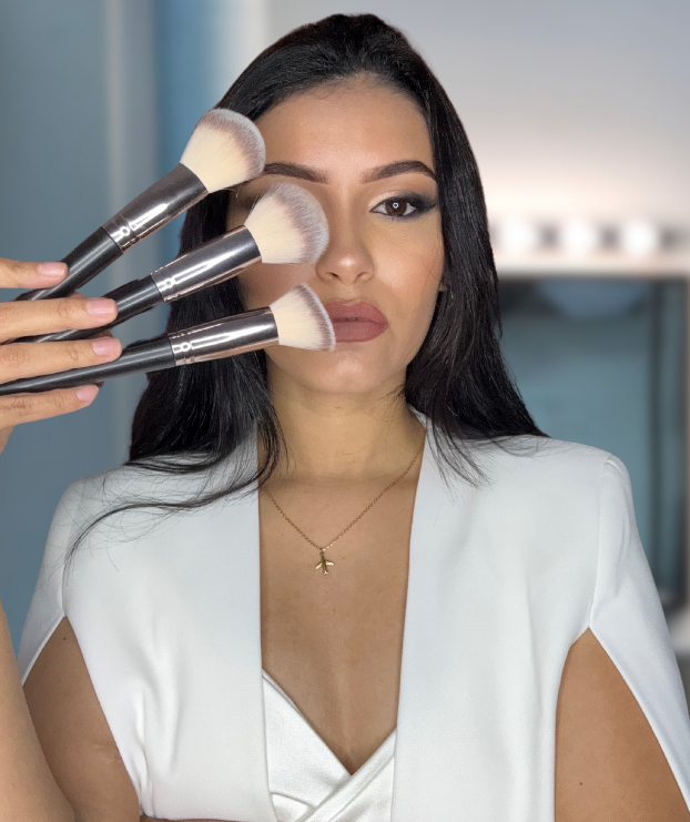 Una mujer con una chaqueta blanca sostiene pinceles de maquillaje frente a su cara.