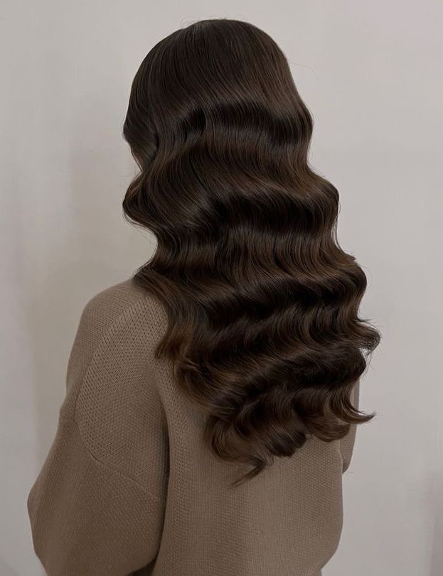 Una mujer con cabello castaño largo y ondulado lleva un suéter marrón.