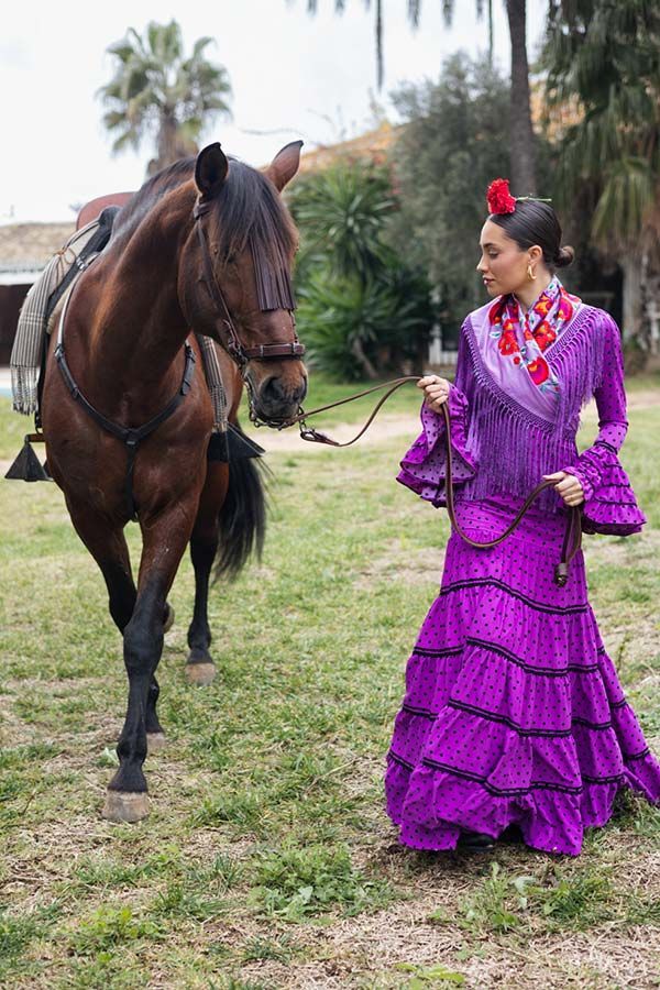 Mujer con un vestido morado brillante con volantes caminando junto a un caballo marrón oscuro sobre la hierba al aire libre.