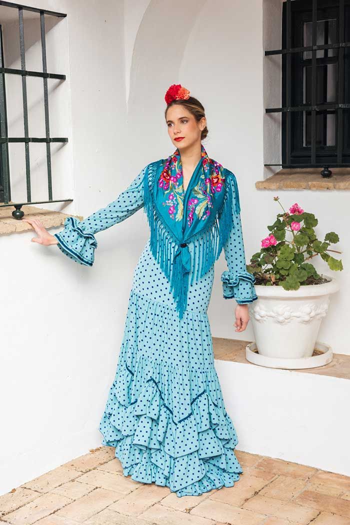 Mujer con un vestido turquesa con volantes posando junto a una pared blanca con una planta en maceta y ventanas negras.