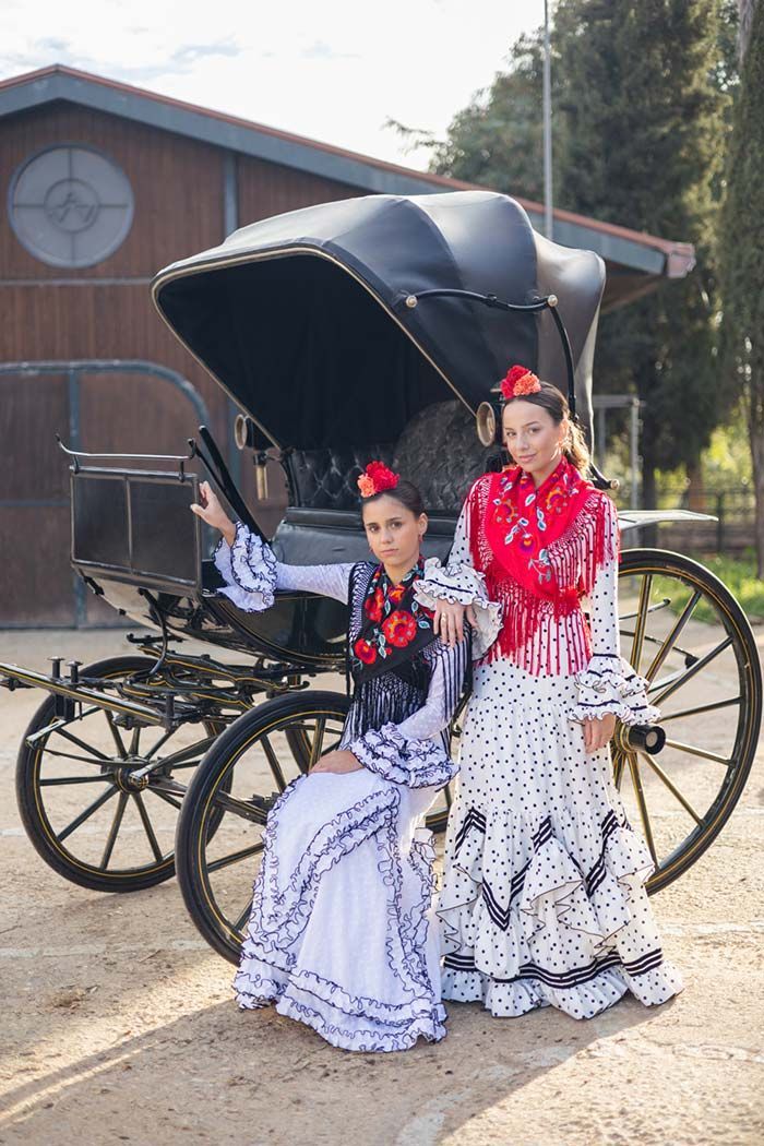 Dos personas con vestidos ornamentados posan junto a un carruaje tirado por caballos al aire libre.
