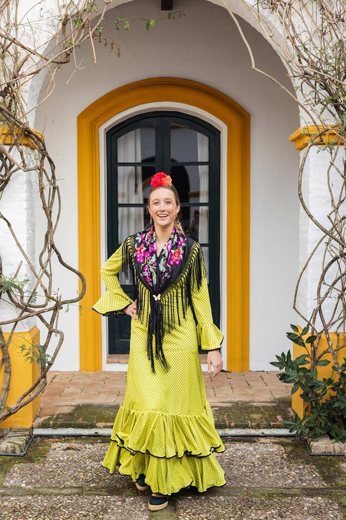 Mujer con un vestido verde lima de pie frente a un arco blanco con una puerta negra y ribete amarillo.