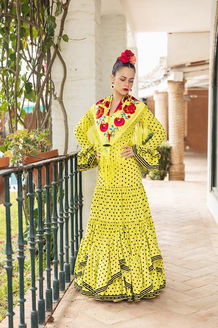 Mujer con un vestido amarillo brillante con lunares negros de pie en un balcón con columnas y barandillas.