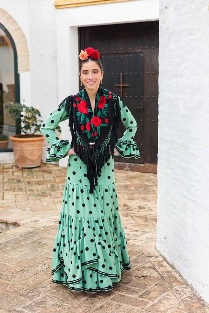 Mujer con un vestido verde menta de lunares de pie en un patio junto a una puerta, con una flor roja en el pelo.
