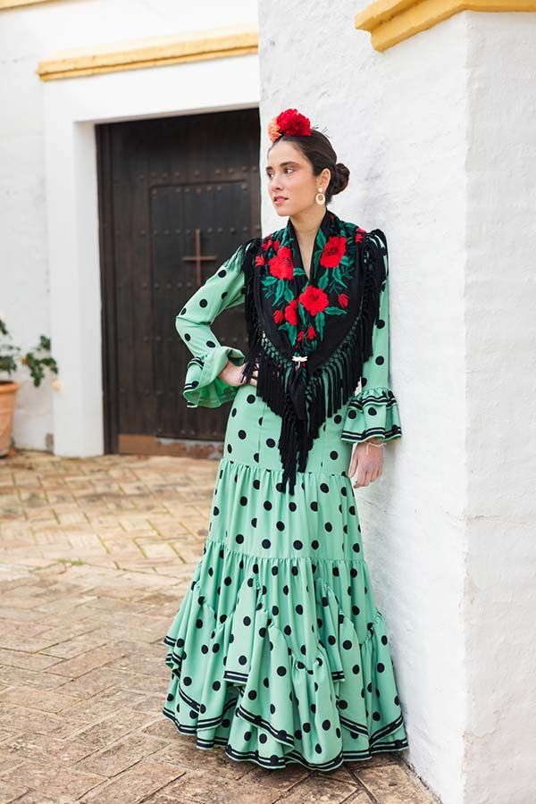 Mujer con un vestido verde menta con volantes y bordados florales rojos, de pie junto a una pared blanca y una puerta.