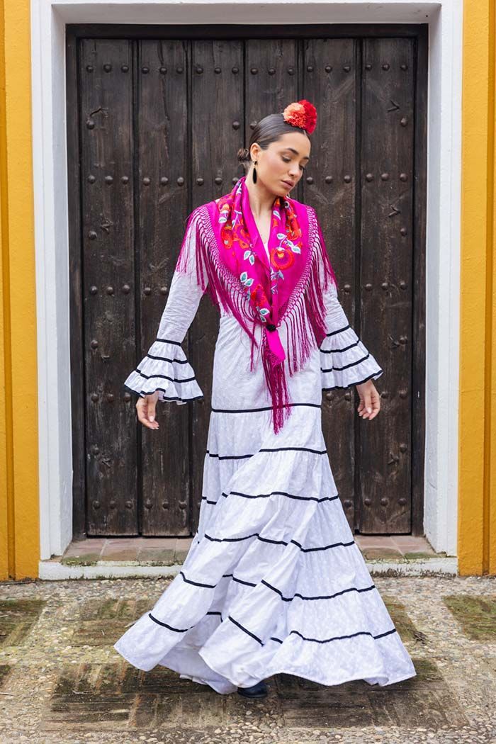 Mujer con un vestido de flamenco blanco y un chal rosa, de pie frente a una puerta de madera oscura.